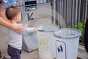 Asian father teach cute little 18 months, 1 year old toddler baby boy child throwing plastic bottle in recycling trash bin at publ