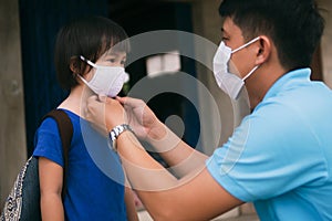 Asian father wearing protective mask puts protective cloth face mask on his little child, schoolkid with backpack is ready to photo