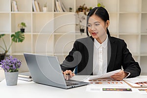 Asian female accountant or banker calculating savings, finance, and economy concepts via laptop. Businesswoman writing charts and