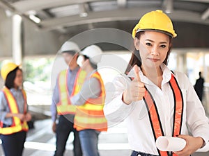 Asian Female architect or engineer holding blue print paper, standing in front of her team smiling , looking at camera and