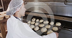 Asian female baker working in bakery kitchen, putting rolls on trays in oven, slow motion