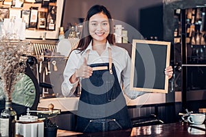 Asian female barista wear jean apron thumbs up at blank blackboard coffee menu at counter bar with smile face,cafe service