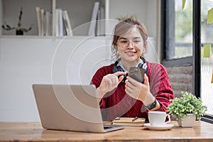 Asian female college student using laptop and phone with headphones while studying. Reading messages and greeting