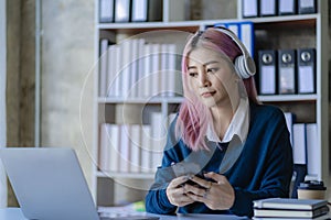 Asian female college student wearing headphones studying online using mobile smartphone app contacting laptop