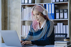 Asian female college student wearing headphones studying online using mobile smartphone app contacting laptop