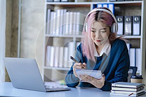Asian female college student wearing headphones studying online using mobile smartphone app contacting laptop