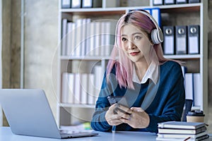 Asian female college student wearing headphones studying online using mobile smartphone app contacting laptop