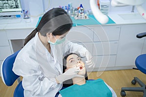 Asian female dentist in lab coat wearing protective face mask to prevent from Covid19. Expert doctor examining tooth for young gir