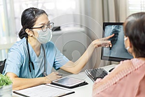 Asian female doctor in medical mask examining and pointing to radiological chest X-ray film on monitor computer with Elderly woman