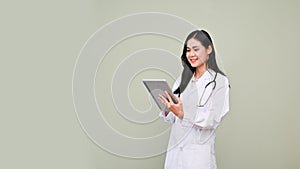 Asian female doctor in uniform using her tablet, standing isolated over grey background