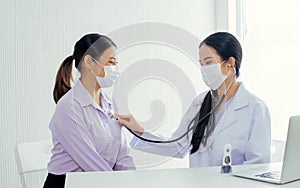 Asian female doctor wearing face mask to protect virus, using stethoscope to listen heart beat of patient getting sick and health