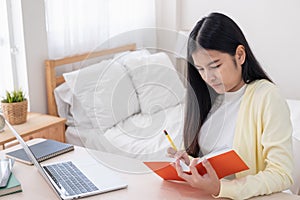 Asian female freelancer reading and jot down note on book while working with laptop on table in bedroom at home.Work at home