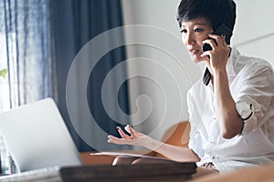 Asian female freelancer sitting on couch talking on phone & working on laptop at home