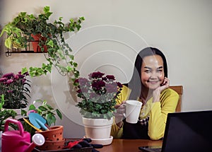 Asian female gardener sitting in her plant workshop drinking coffee and working online on computer laptop  .start up business,