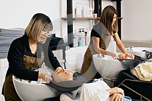 An Asian female hairdresser begins to wash the hair of a woman in a sink in a hair salon.