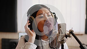 Asian Female Musician Wearing Headphones Singing Song To Microphone In Music Studio