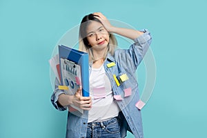 Asian female office worker holding head in hands and holding many document file standing isolated on blue background in studio.