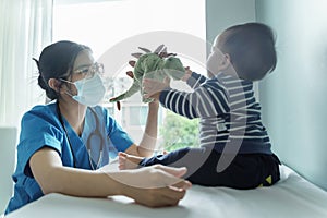 Asian female Pediatrician doctor wearing protective face mask playing dinosaur doll with her little baby boy patient before