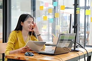 Asian female programmer, wearing a yellow shirt, looking at laptop screen, holding a tablet and pencil. She appeared happy