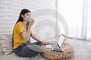 Asian female sit on floor using laptop on wicker stand and drink orange juice in living room at home with relax emotion.Work at