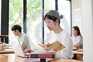 An Asian female student focusing on her work on her laptop and researching information in a book