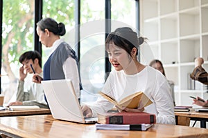 An Asian female student focusing on her work on her laptop and researching information in a book