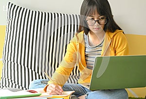 Asian female student wearing glasses is researching with a laptop and taking notes to make a report