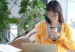 Asian female student wearing glasses is researching with a laptop to report, the other hand holding a coffee cup