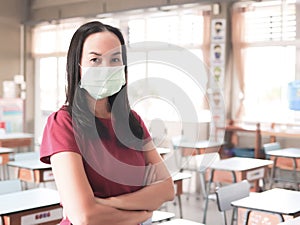 Asian female teacher wearing protective face mask, standing in empty classroom, arms crossed and looking at camera