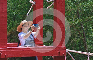 Asian female tourist taking picture with smartphone on red hanging wooden bridge in mangrove forest at nature parkland