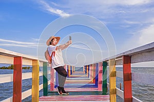 Asian female tourist using smartphone to taking selfie on rainbow wooden bridge at sea viewpoint against blue sky