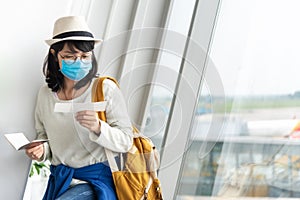 Asian Female tourist wearing protective face mask holding boarding pass waiting for the flight near the window of the airport