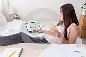 Asian female wear tank top siting and use laptop on bed and drink orange juice in bedroom at home.Work at home concept.work from