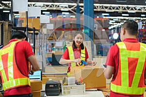 Asian female worker packing cardboard box with tape gun dispenser in warehouse. Thai employee packing goods in large industrial