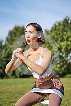Asian fitness girl doing squats in park, using resistance band, stretching yoga rope for workout training on fresh air