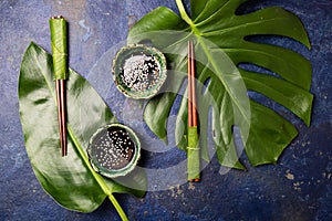 Asian food concept. Chopstick and soysauce soy sauce with white sesame on blue background with tropical leaves . Top