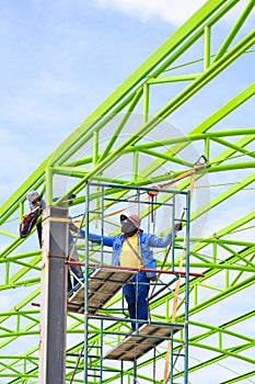 Asian foreman and construction worker on scaffolding are welding metal roof structure of industrial building
