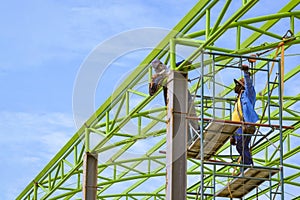 Asian foreman and construction worker on scaffolding are welding metal roof structure of industrial building against blue sky