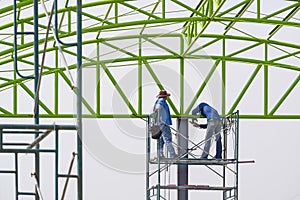 Asian foreman and construction worker on scaffolding are working to build metal roof building structure in construction site