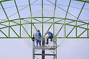 Asian foreman and construction worker on scaffolding are working to build metal roof structure of industrial building in