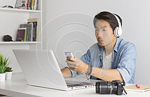 Asian Freelance Videographer Checking Multimedia File by Smartphone with Laptop in Home Office