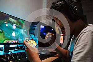 Asian gamer boy screaming while playing video games on smartphone and computer in dark room, wearing headphones and using