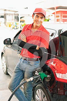 Asian gas station worker man leaning against black car with happiness while green fuel nozzle filling high energy power fuel into