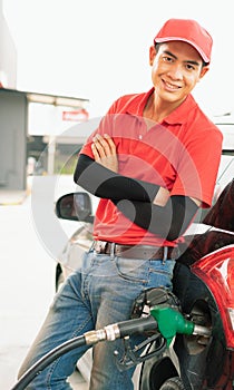 Asian gas station worker man leaning against black car and looking straight with smile while green fuel nozzle filling high energy