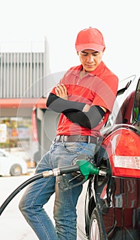 Asian gas station worker man leaning against car and looking to green fuel nozzle which filling high energy power fuel into black