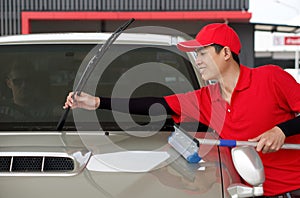 An Asian gas station worker raises the windshield wipers to clean the car windshield. A caucasian driver with sunglass raise