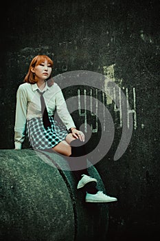An Asian ginger female student, dressed in uniform, sitting on a concrete circle against the backdrop of a gray building wall with