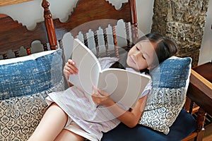 Asian girl child in casual clothes reading a book and lying on bench in the room