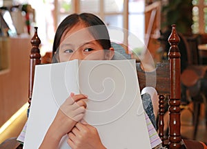 Asian girl child covering faces with white books and looking beside while reading book in library