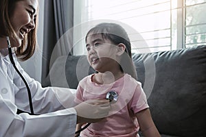 Asian girl child is smiling up and sits on sofa at home during a medical examination while female medical professional is using a